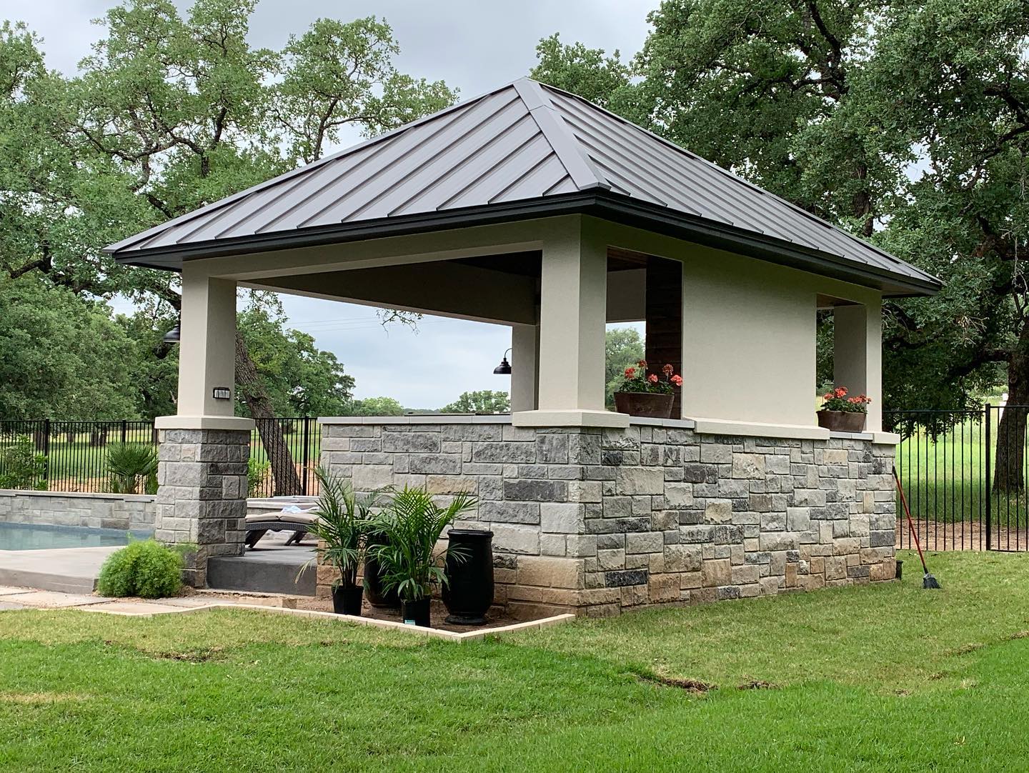 A stone gazebo with a robust metal roof, positioned next to a swimming pool in a lush green yard, providing an elegant outdoor lounging area.