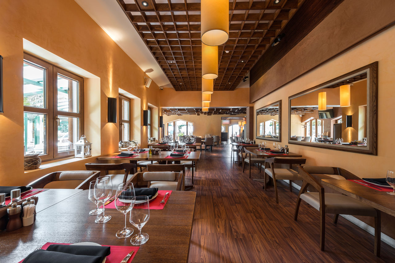 A stylish restaurant interior featuring warm earth tones, wooden furniture, and modern cylindrical lights hanging from a wooden beam ceiling.